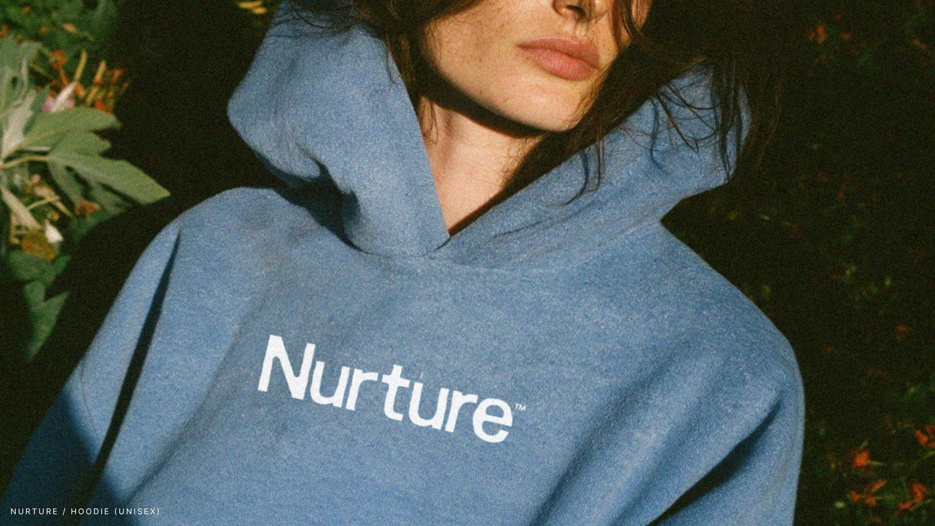 Blue Nurture hoodie with the wordmark across the chest, photographed at night in a garden